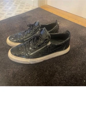 Gieuseppe skor - Säljer ett par svarta sneakers i krokopräglat skinn med snörning och dragkedjor på sidorna. Skorna har vit platt sula och coola detaljer som ger en lyxig vibe. Tillverkade i Italien och passar perfekt för dig som gillar statement-sneakers.