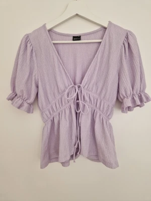 Ljuslila blus med knyt från Gina Tricot - Supersöt ljuslila blus från Gina Tricot i storlek XS. Blusen har puffiga korta ärmar med volangkant, v-ringning och knyt framtill. Tillverkad i ett mjukt, strukturerat tyg som ger en chill och feminin vibe. Perfekt till jeans eller kjol för en söt look.
