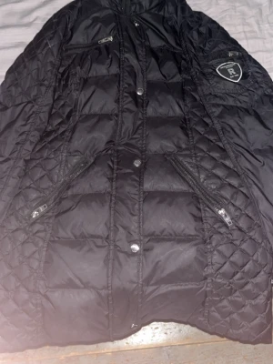 Svart quiltad dunjacka Rockandblue - Säljer en svart quiltad dunjacka från Rockandblue med snygga dragkedjor och logga på ärmen. Jackan har lång ärm, flera fickor med dragkedja och tryckknappar framtill. Materialet är polyester och dun/fjäder, perfekt för kalla dagar.Den är äkta och priset är inte hugget i sten.