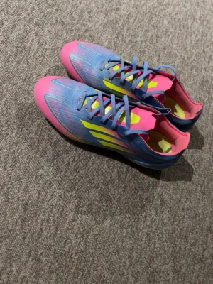 Adidas blå och rosa fotbollsskor - Säljer ett par Adidas F50 elite fotbollsskor i blått och rosa med gula detaljer.Perfekta för spel på gräsplan och även konstgräs. Materialet är syntet för en extrem lätt vikt och bra känsla på bollen. Skorna är i väldigt bra skick. (Priset är ej hugget i sten)