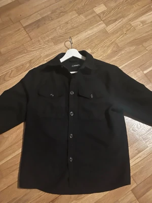  J.Lindeberg overshirt  - Säljer en svart overshirt från J.Lindeberg med klassisk krage, två bröstfickor med knapp och raka ärmar. Jackan har tryckknappar längs hela framsidan och är gjord i ett mjukt ullmaterial. Perfekt för lager på lager och stilren look.