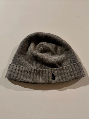 Grå mössa från Polo Ralph Lauren - Snygg grå ribbstickad mössa från Polo Ralph Lauren med klassisk svart logga broderad framtill. Tillverkad i mjukt material som sitter skönt på huvudet och har vikt kant för extra stilpoäng.