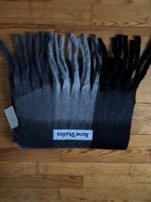 Gråmelerad halsduk från Acne Studios - Säljer en fluffig och mjuk halsduk från Acne Studios i olika nyanser av grått och svart. Halsduken har långa fransar och en stor, vit logotypetikett. Perfekt accessoar för att lyfta din outfit med en stilren touch.