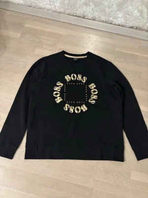 Svart sweatshirt från Hugo Boss - Svart sweatshirt från Hugo Boss med rund hals och tryckt BOSS-logga i gult och vitt framtill. Tröjan har en avslappnad passform och ribbade muddar vid ärmslut och nederkant. Perfekt för en stilren och trendig look.