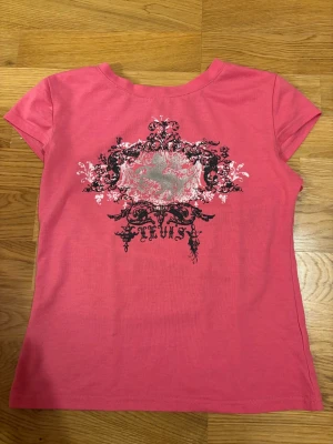 Rosa t-shirt med tryck - Snygg rosa t-shirt med korta ärmar och ett detaljerat tryck i svart, vitt och silver på bröstet. T-shirten har en klassisk passform och är gjord i mjukt material som känns skön mot huden. Perfekt för dig som gillar statement-plagg!