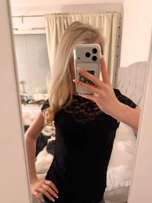 Svart spetsblus med öppen rygg - Säljer en svart blus i spets med korta ärmar och snygg öppen rygg. Blusen har ett feminint spetsmönster över hela och är figurnära i modellen. Perfekt att styla med jeans eller kjol för en trendig look.