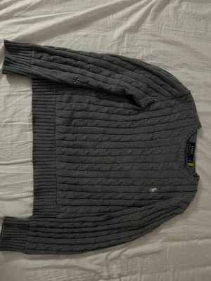 Grå kabelstickad tröja Polo Ralph Lauren - Säljer en mörkgrå kabelstickad tröja från Polo Ralph Lauren med klassisk rund hals och diskret broderad logga på bröstet. Tröjan har långa ärmar och breda ribbade muddar vid ärmslut och nederkant. Perfekt för dig som gillar stilren och tidlös design.