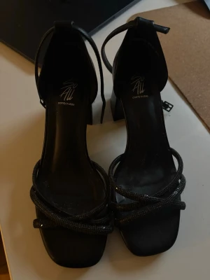 Svarta pumps med remmar från All - Stilrena svarta pumps från All med fyrkantig tå och dekorativa tunna remmar över foten. Skorna har en hög klack och justerbar ankelrem för extra support. Perfekta för dig som vill ha en trendig och elegant look.