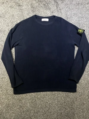 Mörkblå stickad tröja Stone Island - Mörkblå stickad tröja från Stone Island med rund hals och ribbad struktur. Tröjan har lång ärm och den ikoniska Stone Island-loggan på vänster ärm. Perfekt för dig som gillar stilrena och klassiska plagg. Tröjan är i väldigt fint skick och tveka inte att höra av dig:) 