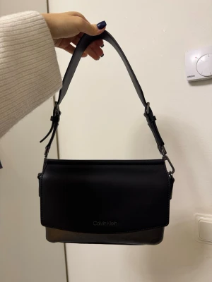 Svart handväska från Calvin Klein - Stilren svart handväska från Calvin Klein med silvrig baksida och justerbart axelband. Väskan har en rektangulär form och är tillverkad i syntetmaterial med diskret logga framtill. Perfekt för dig som gillar minimalistisk design och vill ha en väska som sticker ut.