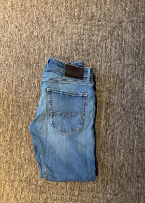 Replay Rocco blå jeans - Säljer ett par Replay Rocco jeans i blå tvätt med med hål på ena benet. Klassisk femficksmodell med raka ben och normal passform. Jeansen har snygga detaljer och är tillverkade i bomull. Perfekt för dig som gillar en avslappnad streetstil.