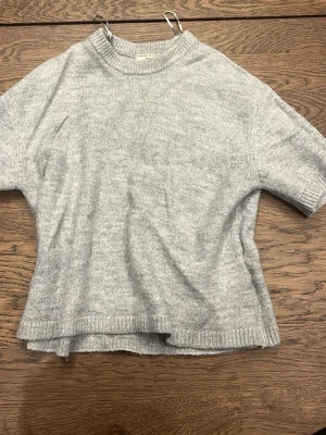 Ljusgrå stickad tröja Gina Tricot - Mysig ljusgrå stickad tröja från Gina Tricot med korta ärmar och ribbade detaljer vid hals, ärmslut och nederkant. Tröjan har en avslappnad passform och är tillverkad i ett mjukt material som passar perfekt till lager på lager.