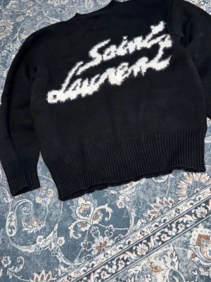 Svart stickad Saint Laurent tröja - Säljer en svart stickad tröja från Saint Laurent med vit, grafisk logga framtill. Tröjan har rund halsringning, ribbade muddar och en avslappnad passform. Perfekt för dig som vill ha en statement piece i garderoben.