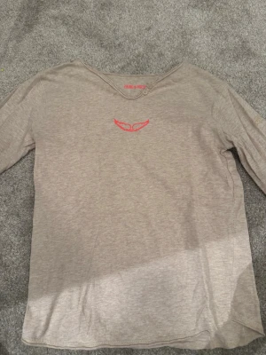 Beige långärmad topp med röd detalj - Säljer en beige långärmad topp från James & Velvet. Toppen har en enkel design med en röd broderad detalj framtill vid halsen. Tillverkad i mjukt bomullsmaterial och har en normal passform. Perfekt för dig som gillar stilrena plagg med en twist.