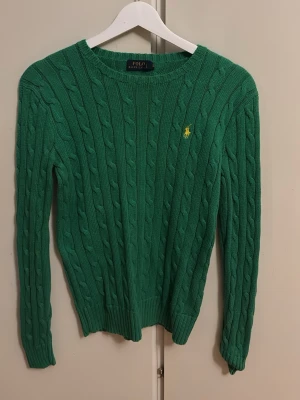 Grön kabelstickad tröja Polo Ralph Lauren - Snygg grön kabelstickad tröja från Polo Ralph Lauren med gul broderad logga på bröstet. Tröjan har rund halsringning, långa ärmar och ribbade muddar. Perfekt för dig som gillar klassisk och tidlös stil.