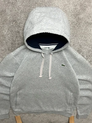 Grå hoodie från Lacoste med huva - Snygg grå hoodie från Lacoste med klassisk huva och dragsnören. Ikoniska gröna krokodilloggan på bröstet. Mjuk bomullsblandning och fickor på sidorna. Perfekt för en chill och stilren look.