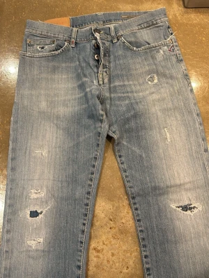 Ljusblå slitna jeans från Dondup - Säljer ett par ljusblå jeans från Dondup med slitningar och hål på benen för en riktigt avslappnad look. Klassisk femficksmodell med raka ben och normal passform. Tillverkade i bomull med snygga detaljer på bakfickorna.