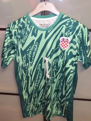 Kroatien målvaktströja Livakovic - Grön målvaktströja från Kroatiens landslag med Livakovic och nummer 1 på ryggen. Tröjan har ett unikt mönster i olika gröna nyanser, rund halsringning och korta ärmar. Kroatiens landslagsemblem på bröstet. Perfekt för fotbollsfans! Den är oanvänd men tvättad. Hör av er om ni har frågor! Priset är ej hugget i sten.