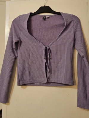 Ljuslila ribbad bolerotopp med knyt - Trendig ljuslila bolerotopp med ribbad struktur och knyt framtill. Toppen har lång ärm och en croppad passform som ger en snygg siluett. Perfekt att styla över ett linne eller klänning för en cool layering-effekt.