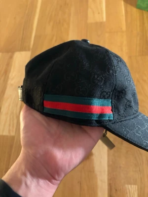 Svart keps från Gucci - Snygg svart keps från Gucci med diskret GG-monogram i tyget och klassiska gröna och röda ränder på sidan. Justerbar rem baktill och böjd skärm. Perfekt accessoar för att lyfta din outfit med lyxig känsla.