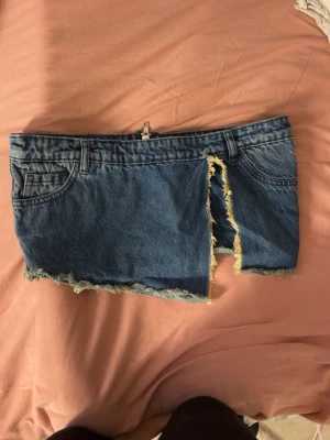 Blå jeanstopp  - Säljer en blå jeanskortkjol i bomull med råa kanter och en markerad slits framtill. Kjolen har bälteshällor, klassiska jeansfickor och dragkedja. Perfekt för dig som gillar edgy denimstil.