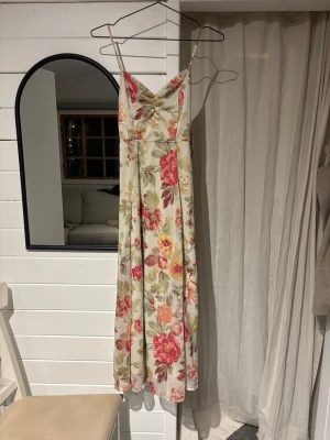 Blommig långklänning med rosett - långklänning med smala axelband och v-ringning. Klänningen har ett blommigt mönster i rosa, rött, gult och grönt på ljus botten samt en stor rosett i ryggen. Perfekt för sommarens alla tillfällen som midsommar 