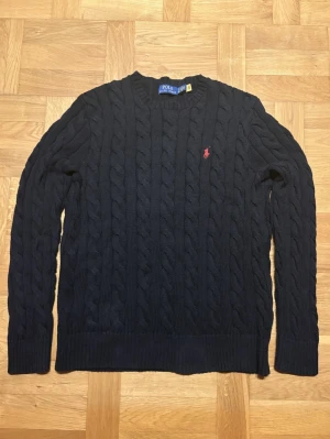 Svart stickad Polo Ralph Lauren tröja  - Säljer en svart stickad tröja från Polo Ralph Lauren med klassiskt kabelstickat mönster och röd broderad logga på bröstet. Tröjan har rund hals och långa ärmar. Perfekt för dig som gillar stilrena och tidlösa plagg. Vid minsta fundering är det bara att höra av sig 