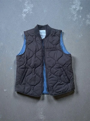 Brun quiltad dunväst från Dan John - Snygg brun dunväst från Dan John  med quiltat mönster och dragkedja framtill. Västen har en klassisk krage och två fickor på sidorna. Perfekt att slänga över en hoodie eller tröja för extra värme och stil. Köpt i lyxbutik/ Italien 