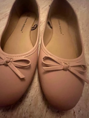 Beiga ballerinaskor med rosett - Snygga beiga ballerinaskor från Guclan Gezzan med rund tå och dekorativ rosett framtill. Skorna har en klassisk och stilren design i syntetmaterial, perfekta för en trendig och enkel look.