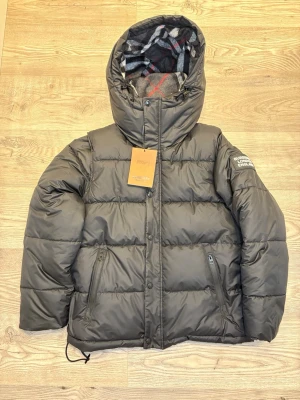 Burberry Jacka - Snygg svart pufferjacka från Burberry med klassiskt rutigt foder i huvan och logga på ärmen. Jackan har dragkedja, två fickor med dragkedja och quiltad design. Perfekt för kalla dagar och riktigt trendig look. Pris kan diskuteras❗️