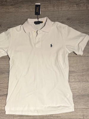 Vit t-shirt Polo Ralph Lauren - Vit t-shirt från Polo Ralph Lauren i bomull med klassisk krage, korta ärmar och blå broderad logga på bröstet. Slim fit-modell som ger en clean och stilren vibe. 