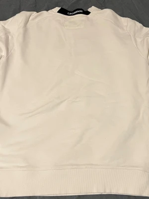 Beige sweatshirt från C.P. Company - Snygg beige sweatshirt från C.P. Company med klassisk rund hals och ribbade muddar. Ärmen har en ikonisk svart linsdetalj och en ficka med dragkedja. Perfekt för en clean och stilren look.