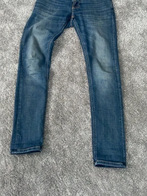 Blå raka jeans från Nudie Jeans - Säljer ett par klassiska blå jeans från Nudie Jeans med raka ben och snygga slitningar. Jeansen har fem fickor, kontrastsömmar och Nudie-logga på bakfickan. Perfekta till vardags för en avslappnad look.