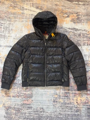 Svart dunjacka med huva Parajumpers - Snygg svart dunjacka från Parajumpers med huva och gul detalj vid halsen. Jackan har quiltad design, dragkedja framtill och två sidofickor med dragkedja. Perfekt för kalla dagar och har en modern, sportig look.