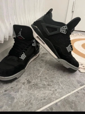Svarta Nike Jordan 4 sneakers - Säljer ett par svarta Nike Jordan 4 sneakers med grå och vita detaljer. Skorna har klassisk snörning, meshpaneler och en platt sula. Ikonisk Jumpman-logga på plösen och cool design som sticker ut. Perfekta för dig som gillar streetwear och sneakers.