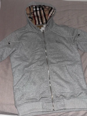 Grå zip hoodie från Burberry - Snygg grå zip hoodie från Burberry med klassiskt rutigt foder i huvan och dragkedja framtill. Hoodien har två stora fickor och ribbade muddar. Perfekt för dig som vill ha en stilren och exklusiv look med en touch av ikoniskt mönster.