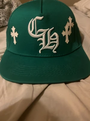 Grön keps med vita kors och brodyr - Säljer en grön keps med platt skärm och vita broderade kors runtom samt stora bokstäver på framsidan. Kepsen har en streetwear-vibe och är tillverkad i bomullstyg. Perfekt för dig som vill sticka ut med en unik accessoar.
