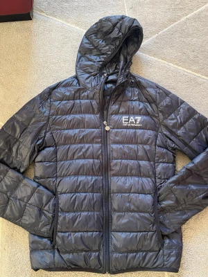  Dunjacka från EA7 Emporio Armani - Snygg svart dunjacka från EA7 Emporio Armani med huva och dragkedja framtill. Jackan har quiltad design och logga på bröstet. Perfekt för kyliga dagar och ger en sportig look. Tillverkad i syntetmaterial med dunfyllning. Priset går att diskutera.