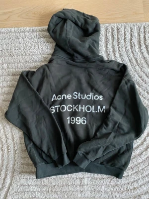 Svart hoodie Acne Studios Stockholm - Cool svart hoodie från Acne Studios med tryck på ryggen: 'Acne Studios STOCKHOLM 1996'. Oversized passform, mjukt material och klassisk huva. Perfekt för dig som gillar streetwear och vill ha en stilren look med tydlig branding.