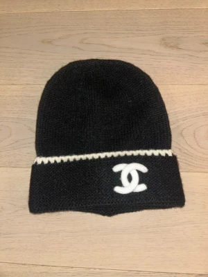 Snygg svart stickad mössa - Snygg svart stickad mössa med vit kant och den klassiska CC-loggan framtill. Mössan har en enkel, rundad form och är perfekt för att lyfta din outfit med en lyxig touch. Materialet är mjukt och värmande, passar perfekt för kyliga dagar.