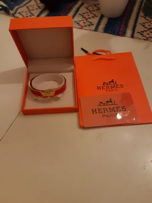 Rött armband från Hermès - Snyggt rött armband från Hermès med guldfärgat spänne och vita detaljer. Kommer i original ask och med Hermès påse. Perfekt accessoar för dig som vill ha något exklusivt och trendigt från ett ikoniskt modehus.