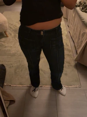 Mörkblå jeans med kontrastsömmar - Säljer ett par mörkblå jeans med vita kontrastsömmar och stora fickor fram. Klassisk femficksmodell med knapp och dragkedja. Snygg passform som sitter bra över höfterna💞💞