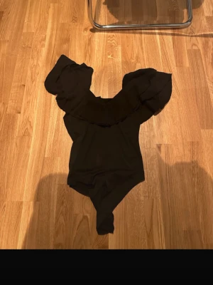 Svart body med volang - Stilren svart body med bred volang runt axlarna som ger en trendig off-shoulder-look. Perfekt för dig som vill ha en snygg och modern stil. Materialet känns mjukt och följsamt.