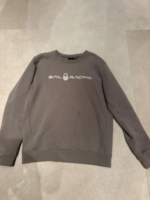Grå sweatshirt från Sail Racing - Snygg grå sweatshirt från Sail Racing med vit logotyp på bröstet. Tröjan har rund halsringning, ribbade muddar och är tillverkad i mjukt material. Perfekt för en avslappnad och sportig stil.