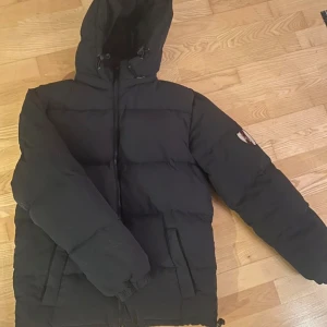 Svart pufferjacka från Kingsland - Svart pufferjacka från Kingsland med huva och dragkedja framtill. Jackan har en quiltad design och en logga på ärmen. Perfekt för kyliga dagar och har en klassisk, stilren look. Hänget på dragkedjan har gått sönder men det än väldigt lätt att dra ipp o ner ändå! Priset kan diskuteras 🥰
