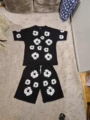 Svart set med vita blommor - Coolt svart set med t-shirt och shorts, båda plaggen har stora vita blommönster tryckta över hela ytan. T-shirten har rund hals och kort ärm, shortsen har resår och vit dragsko i midjan. Perfekt för en avslappnad och trendig stil.