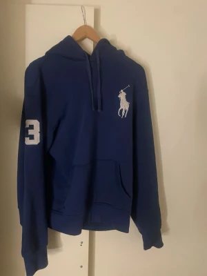 Blå hoodie från Polo Ralph Lauren - Blå hoodie från Polo Ralph Lauren med stor vit logga på bröstet och siffran 3 på ärmen. Klassisk känguruficka och dragsko i huvan. Perfekt för en chill och sportig stil.