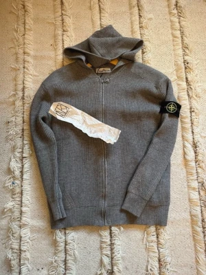 Grå stickad hoodie från Stone Island - Nästan helt ny Snygg grå stickad hoodie från Stone Island med dragkedja och ribbstickad struktur. Klassisk logotyp-patch på ärmen och huva för extra stilpoäng. Perfekt för dig som gillar streetwear och vill ha något bekvämt men ändå trendigt.