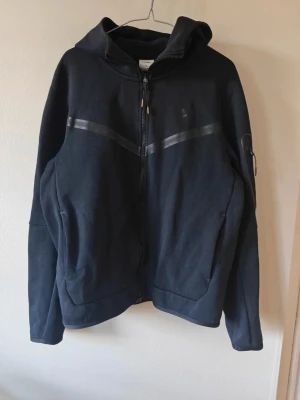 Svart hoodie med dragkedja från Nike. Nike tech - Snygg svart hoodie från Nike med hel dragkedja framtill och två fickor. Hoodien har diskreta svarta detaljer över bröstet och en huva. Perfekt för en avslappnad streetwear-look. Tillverkad i mjukt material för extra komfort. Nike Tech. 