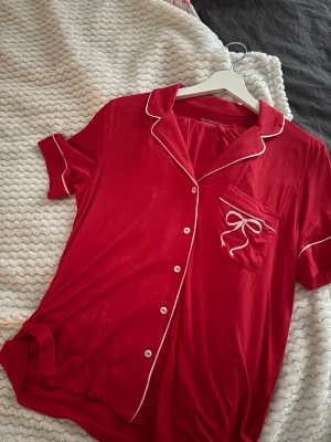 Röd pyjamasskjorta från Victoria's Secret - Snygg röd pyjamasskjorta med korta ärmar från Victoria's Secret. Skjortan har vita knappar, kontrasterande vita kanter och en bröstficka med en söt vit rosett. Perfekt för mysiga kvällar hemma.